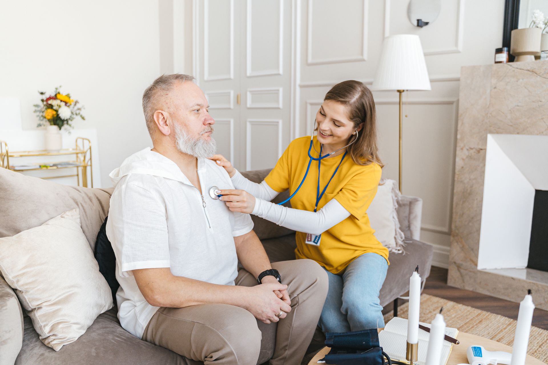 A young healthcare worker in a yellow shirt uses a stethoscope to check the chest of an older man with a white beard, who is sitting on a sofa in a bright living room.