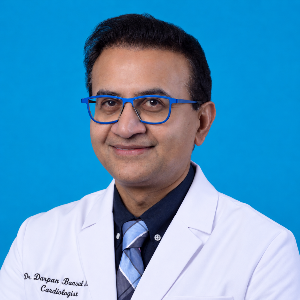 Man with short dark hair wearing blue glasses, a white lab coat, a dark shirt, and a striped tie smiles in front of a solid blue background. The lab coat text reads “Dr. Darpan Bansal, Cardiologist.”.